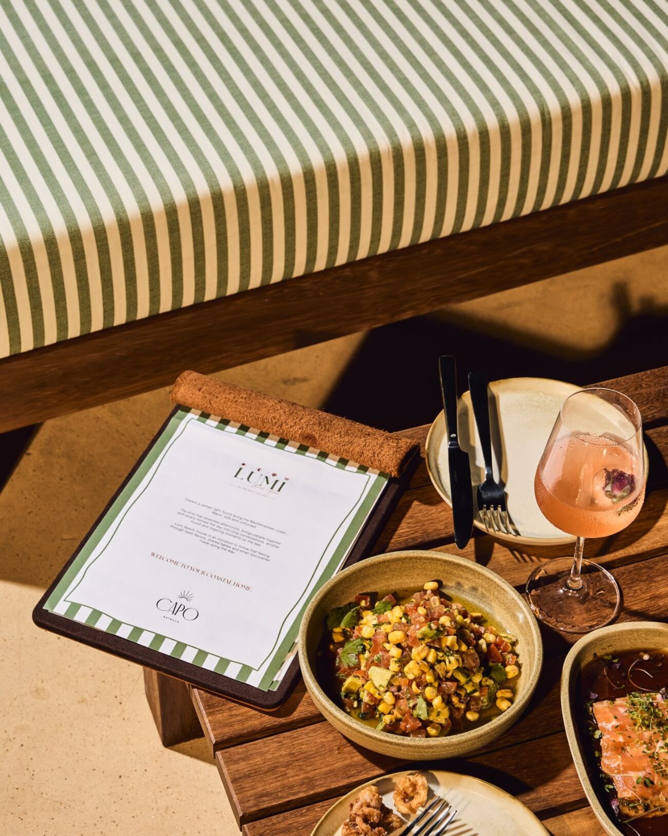 The Lumi menu resting on a striped green and white cushion, beside a rose cocktail and small plates of corn and shrimp on the terrace.