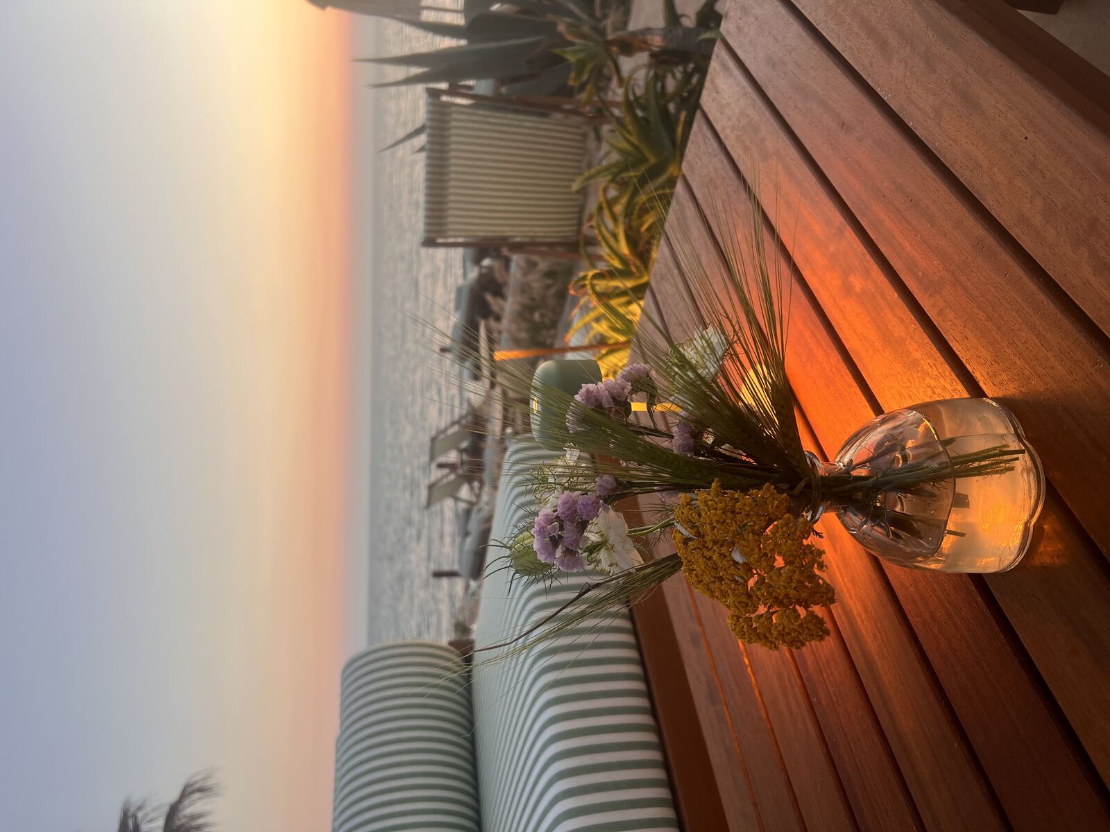A glass vase of wildflowers on a wooden table at golden hour, with striped lounge furniture and the sea behind, at Lumi Beach House.