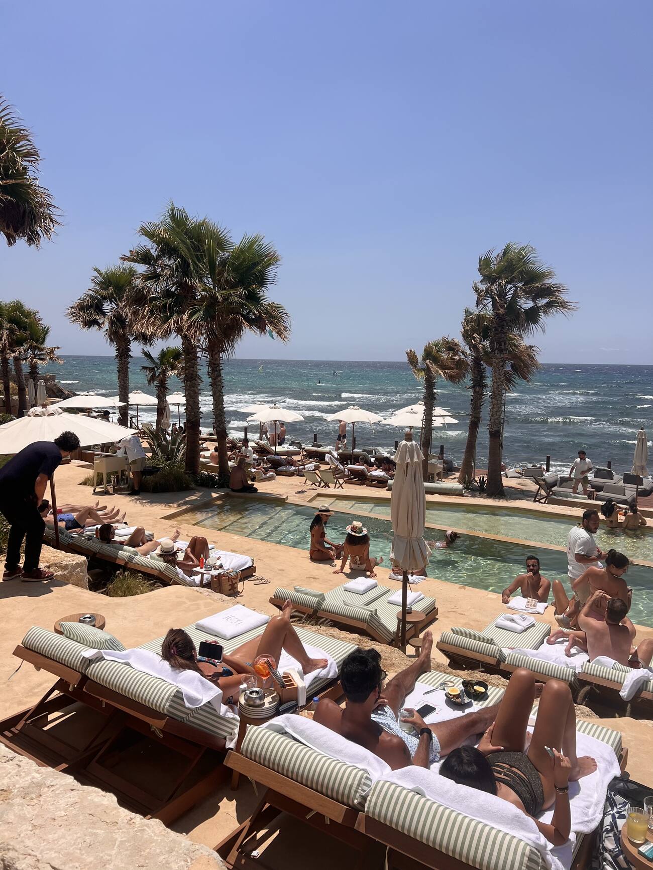 Guests lounging poolside on green and white striped daybeds at Lumi, palm trees and the Mediterranean Sea in the background.